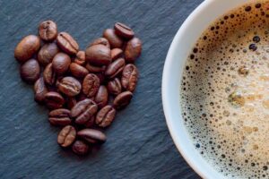 Close-up of heart-shaped coffee beans next to a cup of frothy coffee on a slate surface.